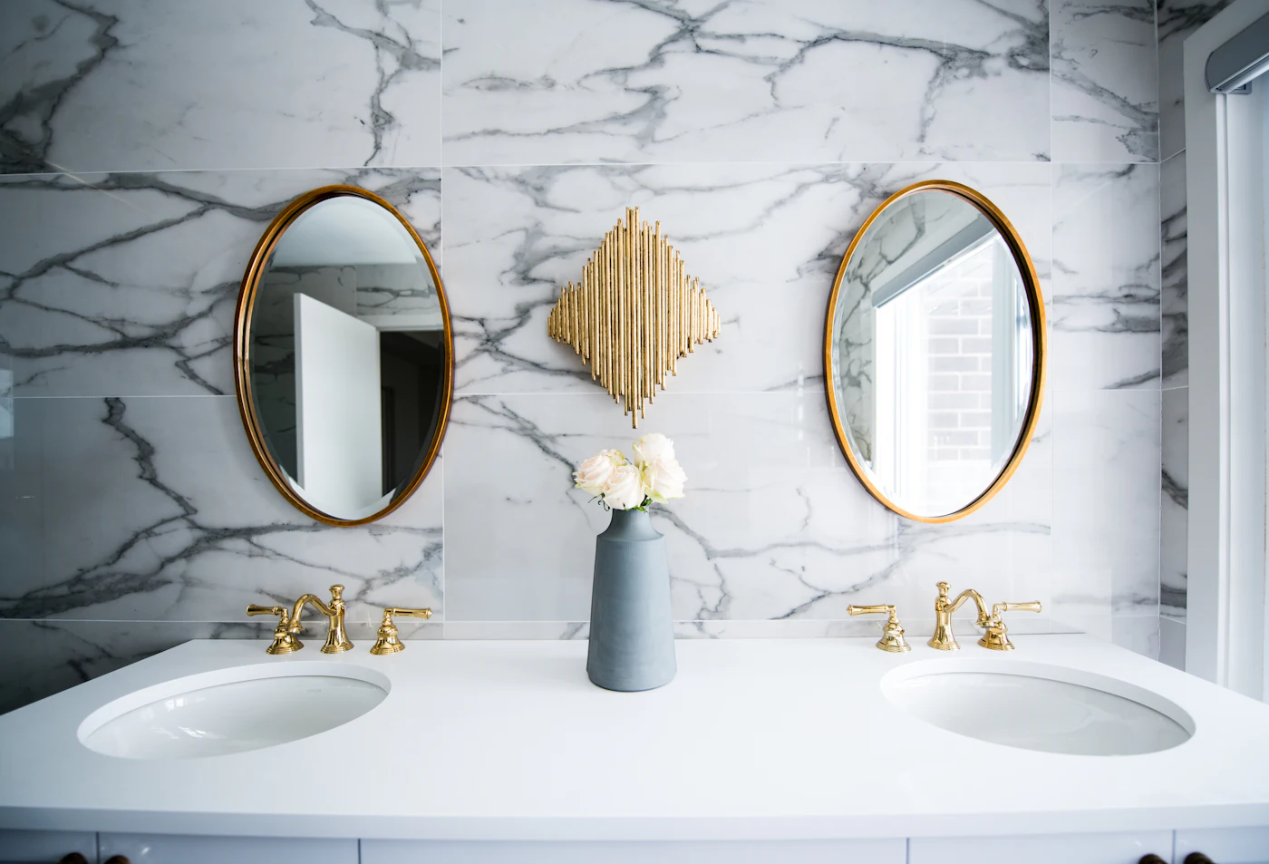 Commercial-style restroom with marble tile wall, double vanity, and brass mirrors